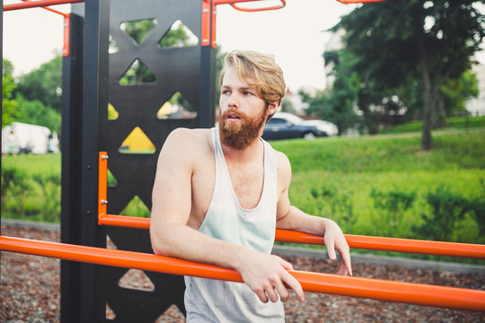 Portrait young bearded man standing on public sports ground and training on parallel bars. Handsome workout athlete. Portrait sporty man with beard, rest after training. Summer workout outdoors