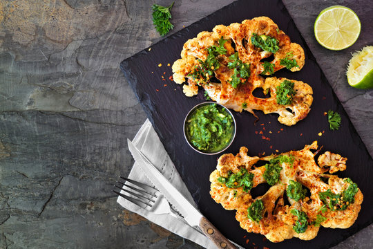 Cauliflower steaks with a cilantro lime sauce. Top view scene on a dark stone background. Healthy eating, plant based meat substitute concept.