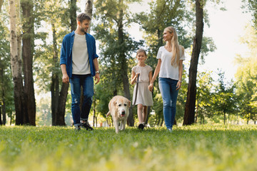 Full length of cute smiling little girl and her parents walking in park with dog