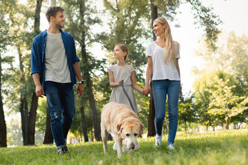 Full length of cute smiling little girl and her parents walking in park with dog and looking on...