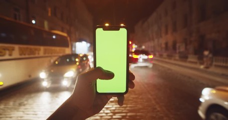 Male hand with vertical chromakey green screen smartphone outdoor. Close-up sociable man using mobile phone at night in busy modern city center. - Powered by Adobe