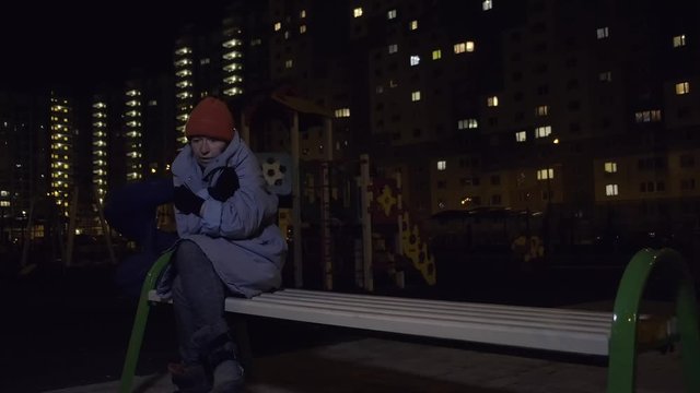 homeless person sits on bench in playground against background of night city. houseless woman is cold and lonely.