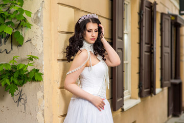 Portrait of Sexy Caucasian Brunette Bride with Tiara Posing Against Old Building with Plenty of Windows. Wearing Beads Necklace and Decorations.