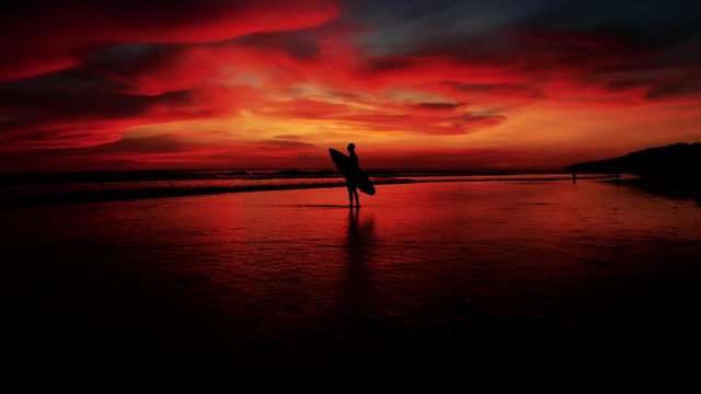 Beautiful aerial of a surfer standing with surfboard in gorgeous dusk sunset red light.