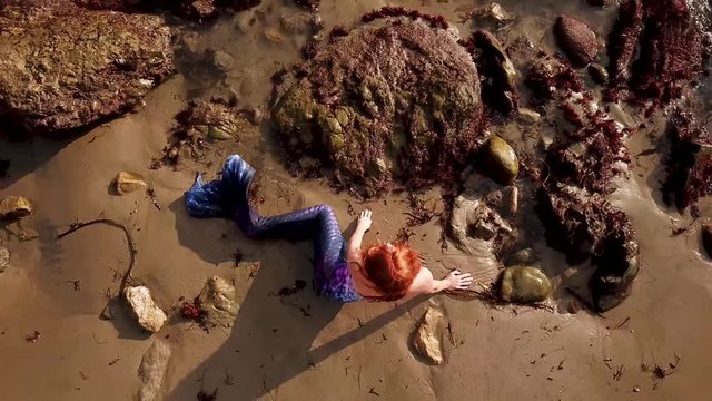 Rising aerial over a fairytale mermaid sitting in ocean waves.