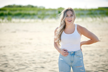 Stunning beautiful woman with blonde hair poses on beach in white tank top and blue jeans
