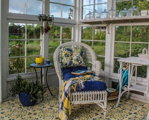 Beautifully decorated interior of a greenhouse/summer house with stenciled floor, white wicker chaise lounge and table, with blue and yellow floral cushions and plaid throw and blue planters