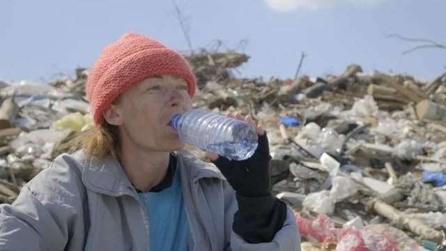 Close-up. homeless woman drinking water from plastic bottle in landfill. bum dwells in the city garbage.