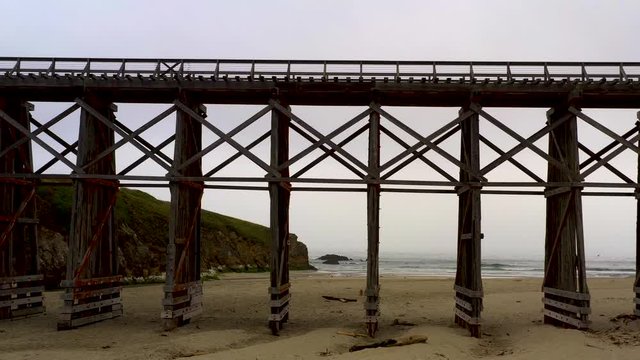 Drone Dolly In from the beach to Pudding Creek Trestle basement, Fort Bragg, California. Fly over sandy beach with ocean waves in a cloudy day.