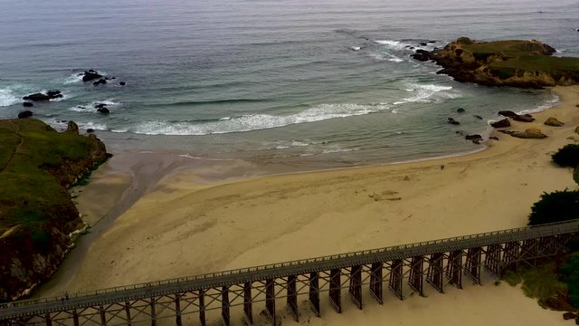 Drone Fly back over ocean waves in sandy beach of Pudding Creek Trestle, part of ten mile beach. Aerial view from seashore to historical bridge in Fort Bragg.