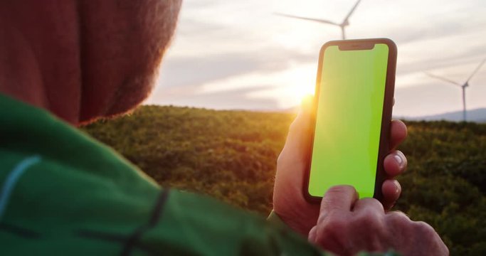 Close-up modern farmer using mobile phone greenscreen at sunset. Beautiful rural landscape with power energy turbines. Ecology farming concept.