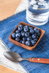 Blueberries in a wood bowl on top of blue and white napkin with a spoon and a cold glass of water