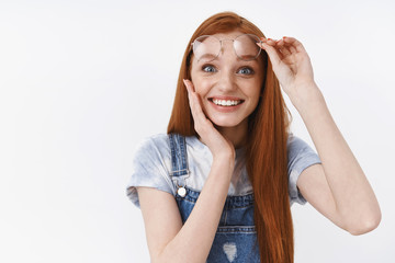 Tender pleased and surprised cute blue-eyed ginger girl smiling toothy amazed, touch cheek delighted satisfied, take-off glasses astonished receive good happy news, stand cheerful white background