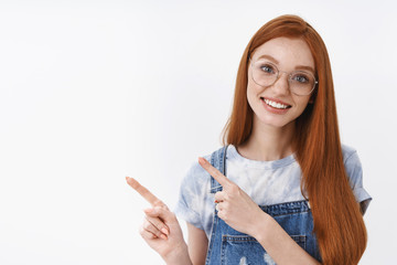 Obraz premium Waist-up silly cheerful redhead female asking question tilt head intrigued, stand white background, pointing left, showing promo, questioned discuss interesting advertisement white background