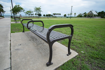 A close up perspective of a dark metal park bench meant to sit on surrounded by the concrete sidewalk surrounded by bright green grass outside on a cloudy day with a pavilion in the background.