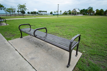 Fototapeta premium A close up perspective of a dark metal park bench meant to sit on surrounded by the concrete sidewalk surrounded by bright green grass outside on a cloudy day with a pavilion in the background.