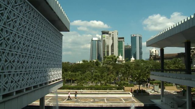 Medium low-angle sunny day still shot of a walking pavement at the National Mosque of Malaysia, compound trees, white masjid domes partially covered by vegetation, and towering closely built city