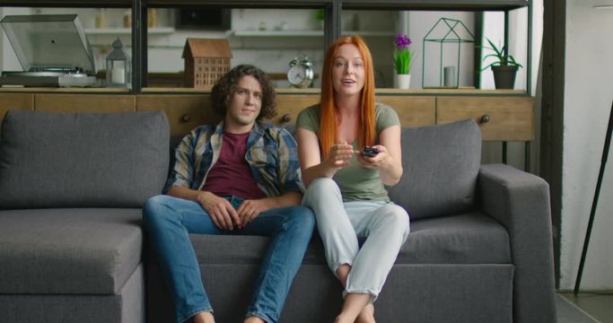 Young couple is watching TV, sitting on sofa in living room, woman is in good mood, watching favorite TV program, show, soap opera, man is bored. Caucasian, white. 4K, shot on RED camera.