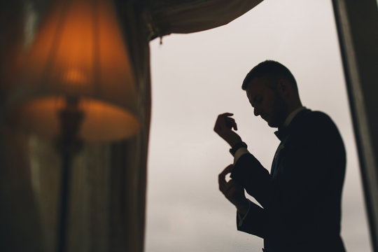 elegant and stylish groom in a blue suit and with a tie butterfly standing in the hotel room near window and getting ready for the wedding
