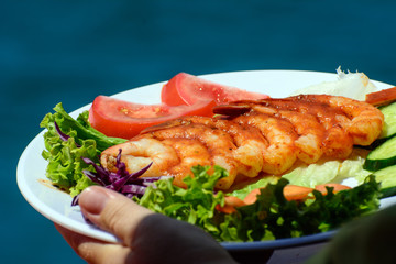 Female hand holds a white plate. Mediterranean shrimps are laid out on a plate. Dish on the background of the sea. Concept - Sea Food