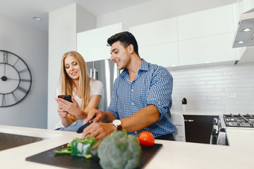 Obraz na płótnie Canvas Man in a kitchen. A handsome man with woman at home. Male and famale at home with vagatables. Blonde with handsome guy prepare salad. Pretty girl use the phone near her boyfriend.