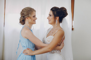 A young and beautiful bride at home standing near window with bridesmaid
