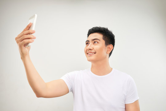 Smiling young asian man making selfie photo on smartphone over white background