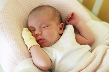 Cute newborn baby with yellow mitten on a hand is sleeping on her bed under beige blanket.