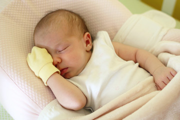 Cute newborn baby with yellow mitten on a hand is sleeping on her bed under beige blanket.
