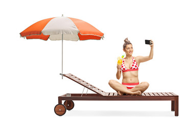 Young woman in bikini sitting on a sunbed under umbrella holding a cocktail and taking a selfie with a phone on a selfie stick