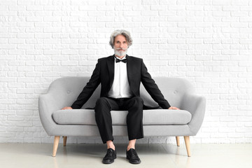Fashionable senior man sitting on sofa near white brick wall