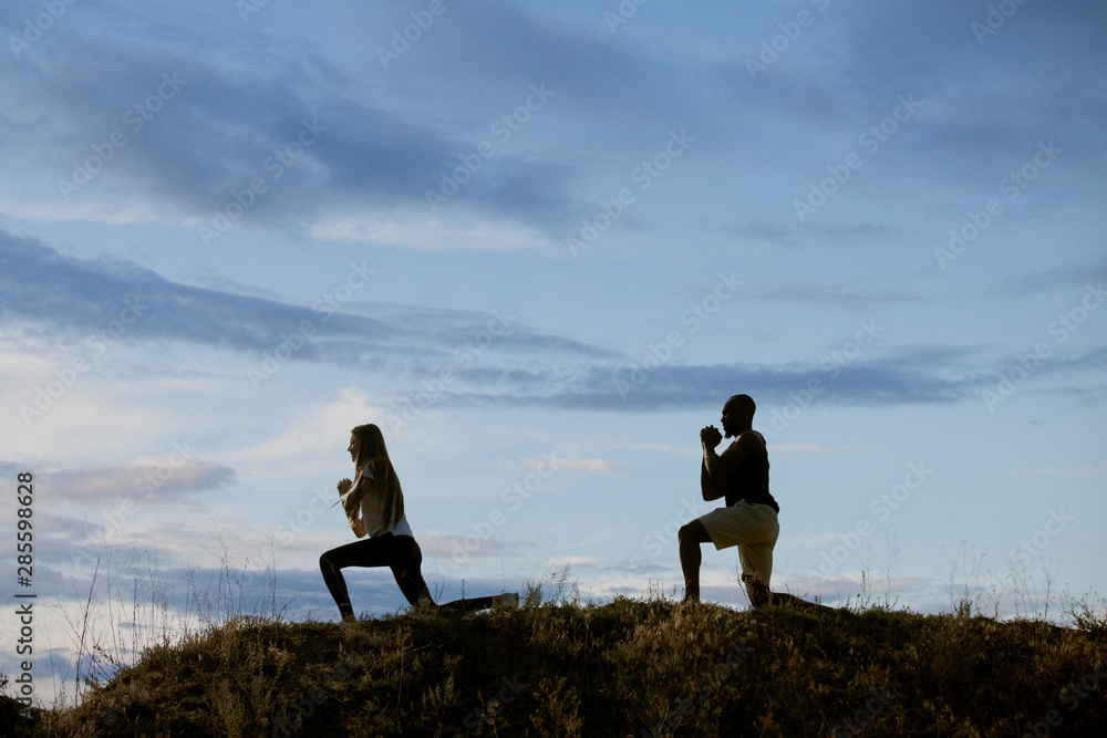 Wall mural Young multiethnic international couple outdoors at the meadow in evening on sky background. African-american man and caucasian woman training and practicing together. Sport, recreation, lifestyle.