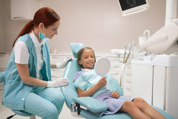 Girl looking at her teeth into mirror while visiting dentist