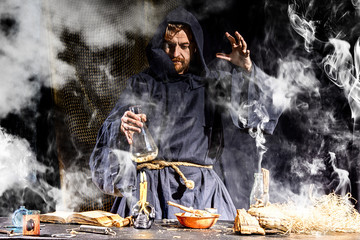 The medieval alchemist make magic ritual at the table in his smoke laboratory.