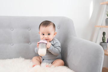 Little cute baby girl sitting in room on sofa drinking milk from bottle and smiling. Happy infant....