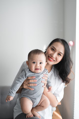 Beautiful young mom is playing with her cute baby and smiling while standing near the window at home