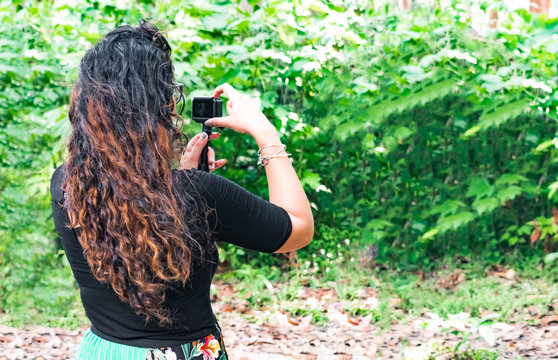 Trendy brunette botanist in her 20s, with curly hairs, is monitoring natural growth of plants during outdoor field work, and is exploring the natural beauty while clicking picture from action camera.