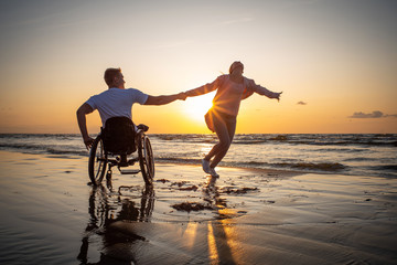 Handicapped man in wheelchair and his girlfriend on a beach at sunset - obrazy, fototapety, plakaty
