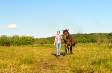 The smiling Caucasian woman is holding her bay horse by the natural halter and leading from a pasture.