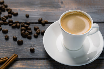 Cup of black coffee on a saucer with cinnamon on a dark wooden table. Coffee beans scattered on the table