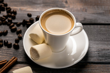Cup of black coffee on a saucer of cinnamon and marshmallow on a dark wooden table. Coffee beans scattered on the table
