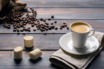 Cup of black coffee and a marshmallow on a linen napkin on a dark wooden table. Coffee beans spill out of the bag on the table