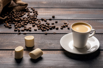 Cup of black coffee and a marshmallow on a dark wooden table. Coffee beans spill out of the bag on the table