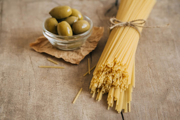 Long yellow raw spaghetti and green olives in glass bowl on wooden background. Copy, empty space for text