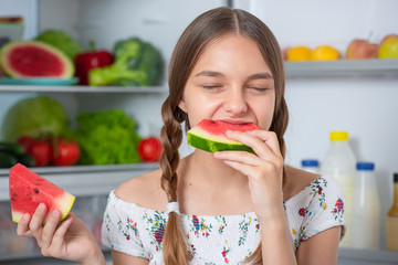 Beautiful young teen girl eating slice ripe red juicy watermelon while standing near open fridge in kitchen at home. Portrait of pretty child choosing food in refrigerator full of healthy products. - 285633689