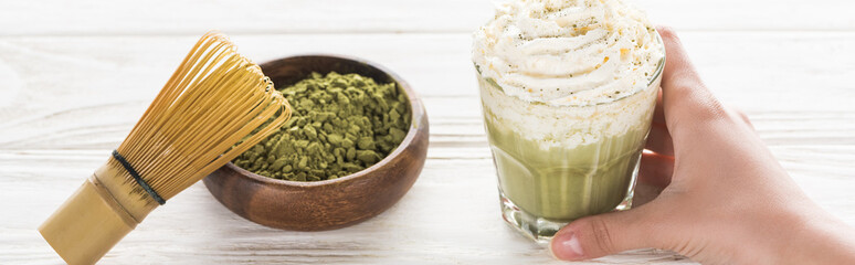 Cropped view of woman holding matcha tea with whipped cream on table with whisk and tea powder