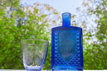 A blue glass water bottle and a glass cup with a blue tint. White and green background. Macro. Water movement.
