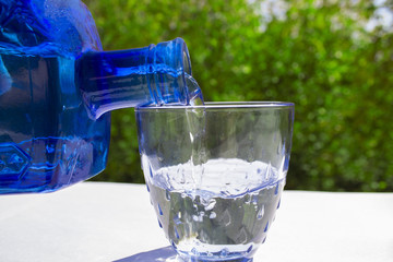 A blue glass water bottle and a glass cup with a blue tint. White and green background. Macro. Water movement.