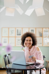 Beautiful mixed race woman in pink turtleneck sweater sitting in pastry shop and using laptop for on line shopping.