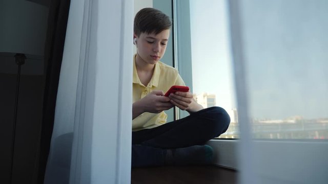 A young handsome man sits at the window with non-wired headphones behind the curtain and looks at the screen of his smartphone